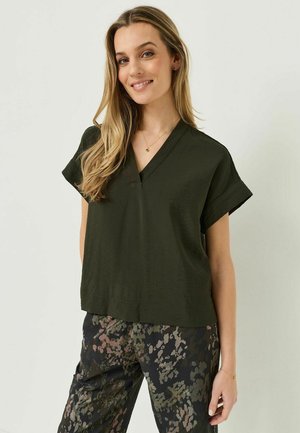 Donna sorridente con capelli biondi lunghi che indossa una maglia verde scuro a scollo a V e pantaloni con motivo floreale, in piedi davanti a uno sfondo chiaro.