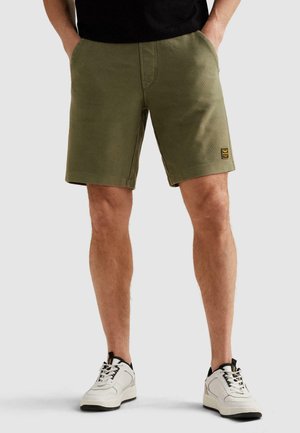 Mann trägt olivgrüne Shorts mit Seitentaschen und weißen Sneakern, steht mit den Händen in den Taschen vor einfachem Hintergrund.