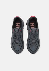 Graue Sportschuhe aus Mesh und Synthetikmaterial, runde Zehenpartie, schwarze Gummisohle und rote Akzente an Ferse und Schnürsenkeln.