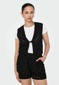 Must mustriline vest, millede has niidud mustrist, kinnitatud ees, valge T-särgiga; paaris mustade pitsist lühikeste pükstega, millel on sarnane disain.