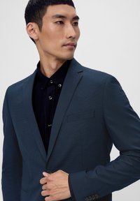 Dunkelblauer Blazer mit strukturiertem Muster, einfacher Knopfverschluss, Reverskragen und einer Brusttasche, getragen über einem schwarzen Hemd mit Knöpfen.