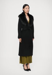 Melns apkārtējais mētelis ar mākslīgās kažokādas apkakli, garām piedurknēm un jostu, valkāts virs zaļas kroku kleitas un melniem augstpapēžu apaviem.