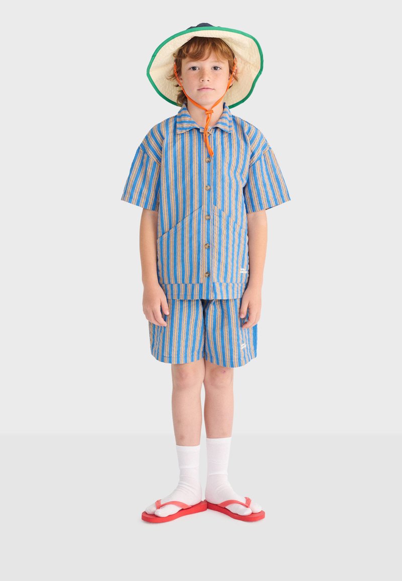 Barn iført stribet blå og orange kortærmet skjorte og shorts, solhat med hakestropp, hvide sokker og røde klipklappere, står stille.