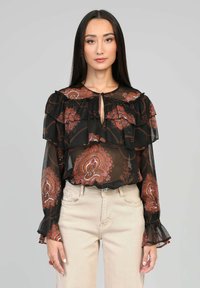 Blouse noire transparente avec des volants, motifs floraux en bordeaux et rose, accentuée d'un décolleté en trou de serrure et de poignets à manches évasées.