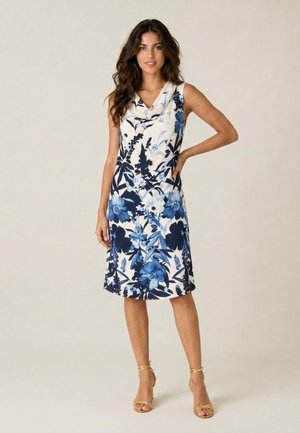 Donna in piedi che indossa un vestito bianco senza maniche, lungo fino al ginocchio con motivi floreali blu e sandali dorati con cinturini e tacchi alti.