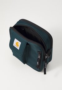 Bolsa de tela en color verde azulado con dos cremalleras negras, bolsillo frontal y logo de Carhartt. Forma cuadrada compacta y textura duradera para versatilidad y funcionalidad.