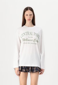 Bijela majica dugih rukava s zelenim natpisom "CENTRAL PARK Wellness Cake" s okruglim izrezom i ležernim krojem.