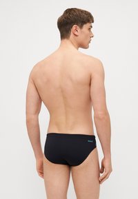 Sorte svømmedrakter laget av elastisk materiale, med et enkelt design, lavt liv og liten logo i blått på siden.