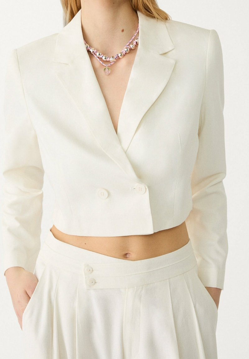 Blazer blanco cortado con mangas largas, frente de doble botonadura y textura suave; combinado con pantalones de talle alto con pliegues que presentan detalle de botones.