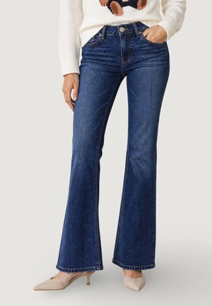 Femme portant un jean évasé bleu foncé et des chaussures beige à talon kitten, une main dans la poche, pull blanc partiellement visible.