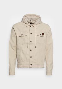 Beige denimová bunda s připojenou kapucí, patkovými knoflíky, dvěma náprsními kapsami a třepeným lemem. Obsahuje malou nášivku na levé straně hrudi.