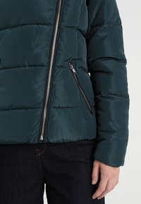 Vadderad, mörkgrön jacka med diagonala sömmar, fullständig dragkedja framtill och detaljer med dragkedjefickor. Slät, glänsande yta.