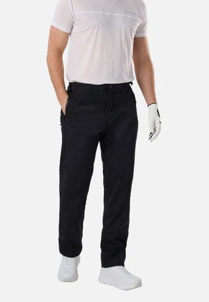 Mężczyzna w białej koszuli z krótkim rękawem, czarnych spodniach z kieszeniami na zamek, białej rękawiczce na prawej ręce i białych tenisówkach, stojący na białym tle.