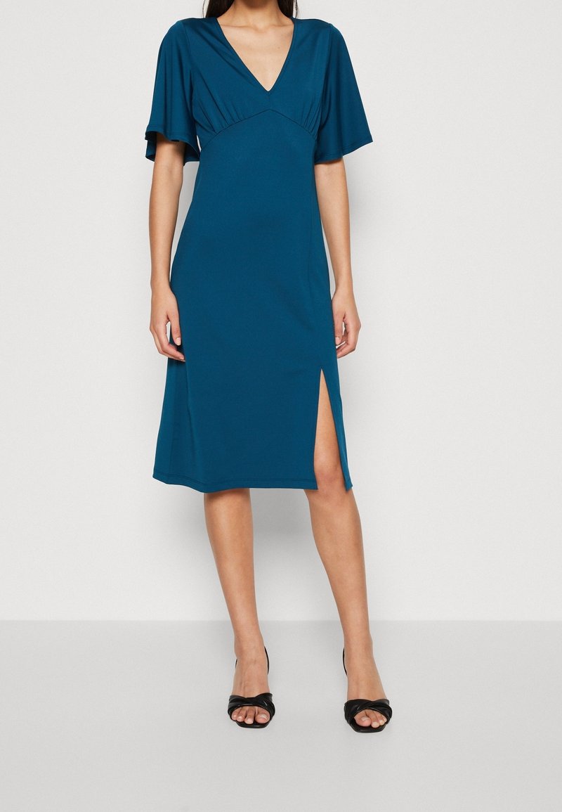 Robe bleu sarcelle longueur genou avec encolure en V, manches courtes évasées et fente latérale. Confectionnée dans un tissu doux à la texture lisse.