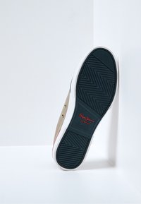 Zapatillas de lona beige con suela de goma blanca, presentando una suela azul texturizada y un acento rojo en el talón, de la marca "Pepe Jeans London."