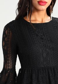 Kvinna i en svart spetsklänning med intrikata blommönster, röd läppstift, långt mörkt hår och guldhängsörhängen med barpendlar.