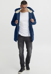 Jūras spēku parka jakas ar mākslīgās kažokādas oderi kapuci, rāvējslēdzēja atvēršanu un stringu jostu; valkāta virs baltas krekliem un saskaņota ar pelēkiem džinsiem.