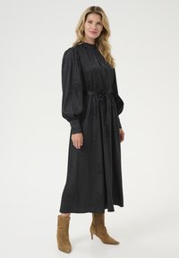 Musta pitkähihaisen mekko, jossa on teksturoidut kukka-ompelut, vyötärö kireällä kangashihnalla, yhdistettynä vaaleisiin nilkkureihin.