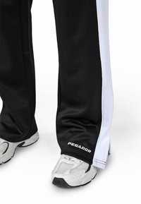 Calças de treino pretas com riscas laterais brancas, com um corte straight e logótipo "PEGADOR" na bainha. Usadas com ténis brancos de desporto.