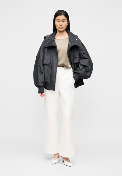 Juodas per didelis švarkas su didelėmis kišenėmis, smėlio spalvos palaidinė, baltos platėjančios kelnės ir smailaus priekio baltos aukštakulniai, ant paprasto fono.