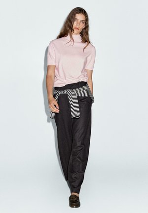 Giovane donna con capelli castani sciolti, che indossa un dolcevita a maniche corte rosa pallido, pantaloni neri, un maglione a righe legato in vita e mocassini scuri.