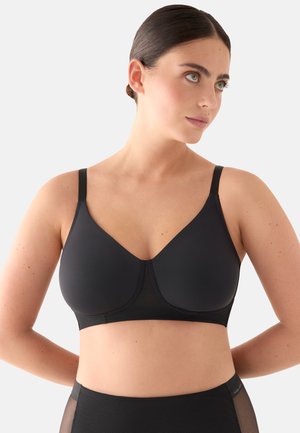 Mujer de cabello oscuro con un sujetador negro y ropa interior negra de talle alto, mirando hacia un lado contra un fondo claro y sencillo.