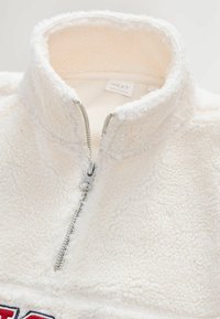 Fleece félzip pulóver törtfehér színben, ezüst cipzárral, texturált anyaggal, és egy márka címkével a gallér belső oldalán.
