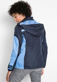 Giacca a vento navy e azzurro chiaro con cappuccio, caratterizzata da pannelli laterali a contrasto e polsini a righe, realizzata in un materiale liscio e leggero.