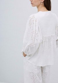Blusa bianca in cotone con maniche a sbuffo, caratterizzata da motivi geometrici a taglio e una vita aderente. Abbinata a pantaloni coordinati con lo stesso motivo.
