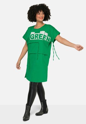 Robe verte surdimensionnée avec le texte "GREEN", manches courtes et taille à cordon. Associée à des leggings noirs et des bottes à cheville cloutées.