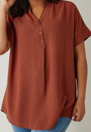 Femme portant un chemisier texturé à manches courtes de couleur rouille avec trois boutons à l'avant, associé à un jean bleu clair.