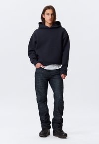 Sweatshirt à capuche bleu marine sombre avec une poche avant, associé à un jean texturé sombre et des chaussures noires pour un look décontracté.