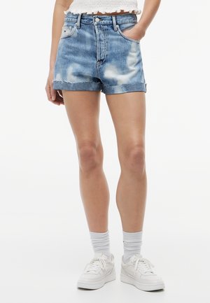 Nainen pukeutuneena sinisiin tie dye -farkkushortseihin, joissa käännetyt lahkeet, valkoiset tennarit, valkoiset sukat ja valkoinen lyhyt toppi, kädet osittain taskuissa.
