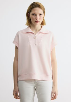 Νεαρή γυναίκα με ανοιχτόχρωμα μαλλιά, ντυμένη με ανοιχτό ροζ μπλουζάκι με κοντά μανίκια και φερμουάρ και λευκό παντελόνι, στραμμένη προς τα εμπρός με ουδέτερη έκφραση.