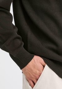 Blizu prikaz tamnog pulovera osobe s rebrastim rukavima, prikazujući ruku opušteno smeštenu u džep svijetlih hlača.