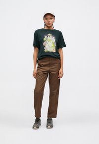 T-shirt graphique vert avec motif floral, pantalon marron et chaussures grises. Le mannequin porte une casquette claire ; style simple et décontracté.