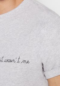 T-shirt en coton gris avec manches courtes, encolure ronde, et texte brodé noir "ce n'était pas moi" sur le devant.