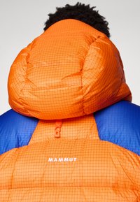 Chaqueta acolchada con un estampado a cuadros naranja y un panel azul. Incluye una gran capucha acolchada y un logo en el pecho.
