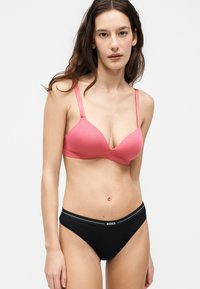 Femme portant un soutien-gorge rose et un sous-vêtement noir de la marque BOSS, debout devant un fond clair uni avec une expression neutre.