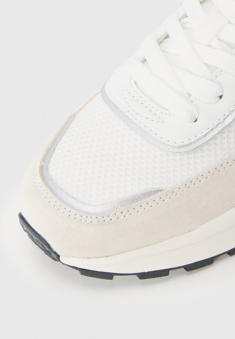 Witte mesh en suède sneaker met zilveren accenten, ronde neus, platte veters en een zwarte rubberen zool met een gestructureerd patroon.
