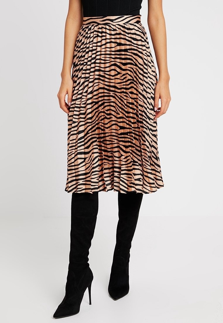 Femme portant une jupe midi plissée avec un imprimé zèbre beige et noir, associée à des bottes noires hautes jusqu'au genou à talons pointus.