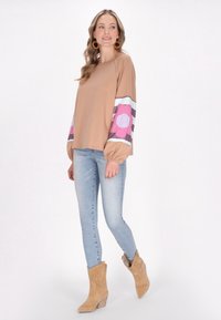 Långärmad beige topp med blommönstrade ärmar i rosa och lila, kombinerad med ljusblå jeans och tan-färgade, avslappnade ankelstövlar.