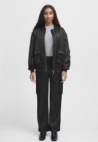 Jachetă bomber din satin negru cu două buzunare frontale, cămașă gri cu dungi, pantaloni cargo negri, slim fit, și pantofi negri lucioși.