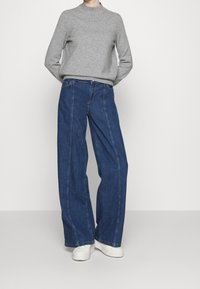 Pull en tricot gris associé à un jean en denim bleu foncé, taille haute et ample. Le jean présente des coutures visibles et des poches avant. Baskets blanches.