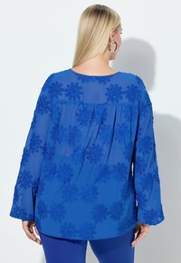 Blå bluse med lange ærmer, gennemsigtigt stof og tekstureret floral broderi. Har en rund halsudskæring og et rynket bagpå design.