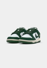 Une paire de baskets Nike blanches et vertes foncées avec des lacets et le logo swoosh, disposées côte à côte sur un fond blanc.