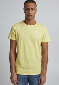 Gul bomull t-skjorte med rund hals, korte ermer og et lite hvitt grafisk design med palmer på venstre bryst.