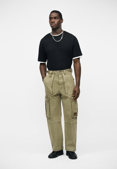 Άνδρας στέκεται με τα χέρια στις τσέπες, φοράει μαύρο μπλουζάκι, ασημένια αλυσίδα, μπεζ παντελόνι cargo και μαύρα παπούτσια, σε λευκό φόντο.
