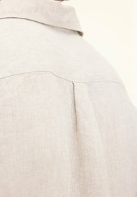 Camicia in lino beige chiaro con colletto e dettagli di piega sul retro. Il tessuto sembra avere una texture e essere traspirante, adatto per il clima caldo.