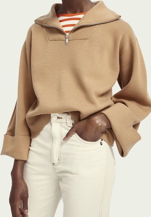 Personne portant un pull beige à demi-fermeture éclair sur une chemise rayée orange et blanche, une main dans la poche d’un pantalon blanc taille haute.